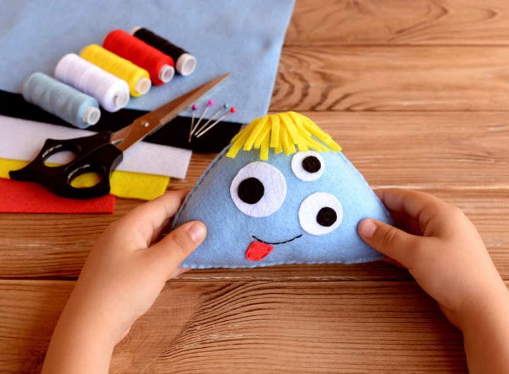 child's hands hold felt craft funny monster face with three googly eyes, thread and felt in bacground